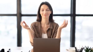 A woman at a desk raises her hands in excitement while working on her laptop.